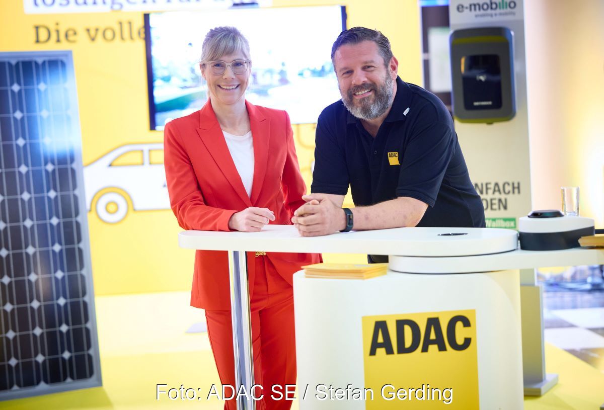 Heike Löffler von Lichtblick und Sascha Coccorullo vom ADAC an einem Stand des ADAC mit PV-Modulen im Hintergrund.
